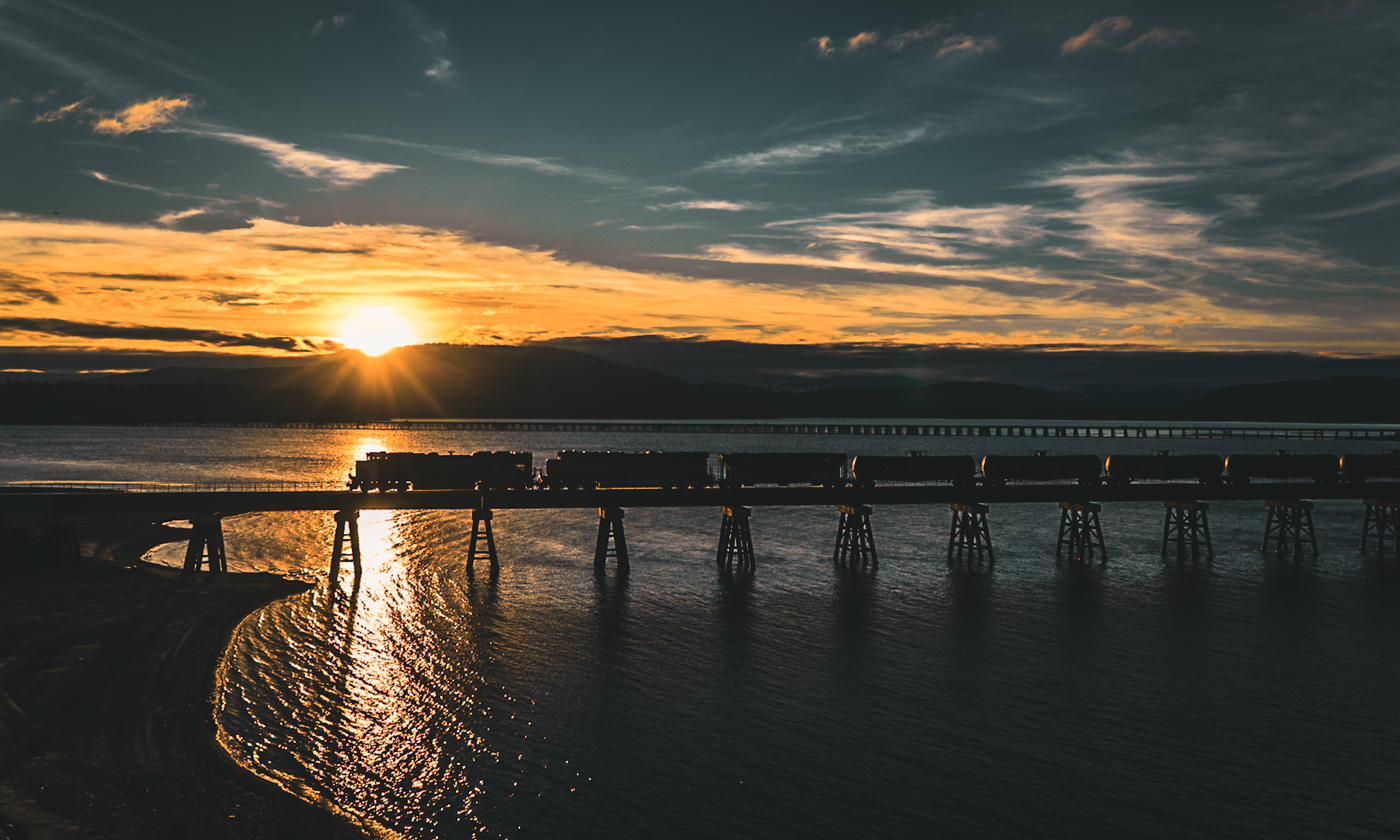 Train crossing a river trestle at golden hour, silhouetted against a blazing western sky