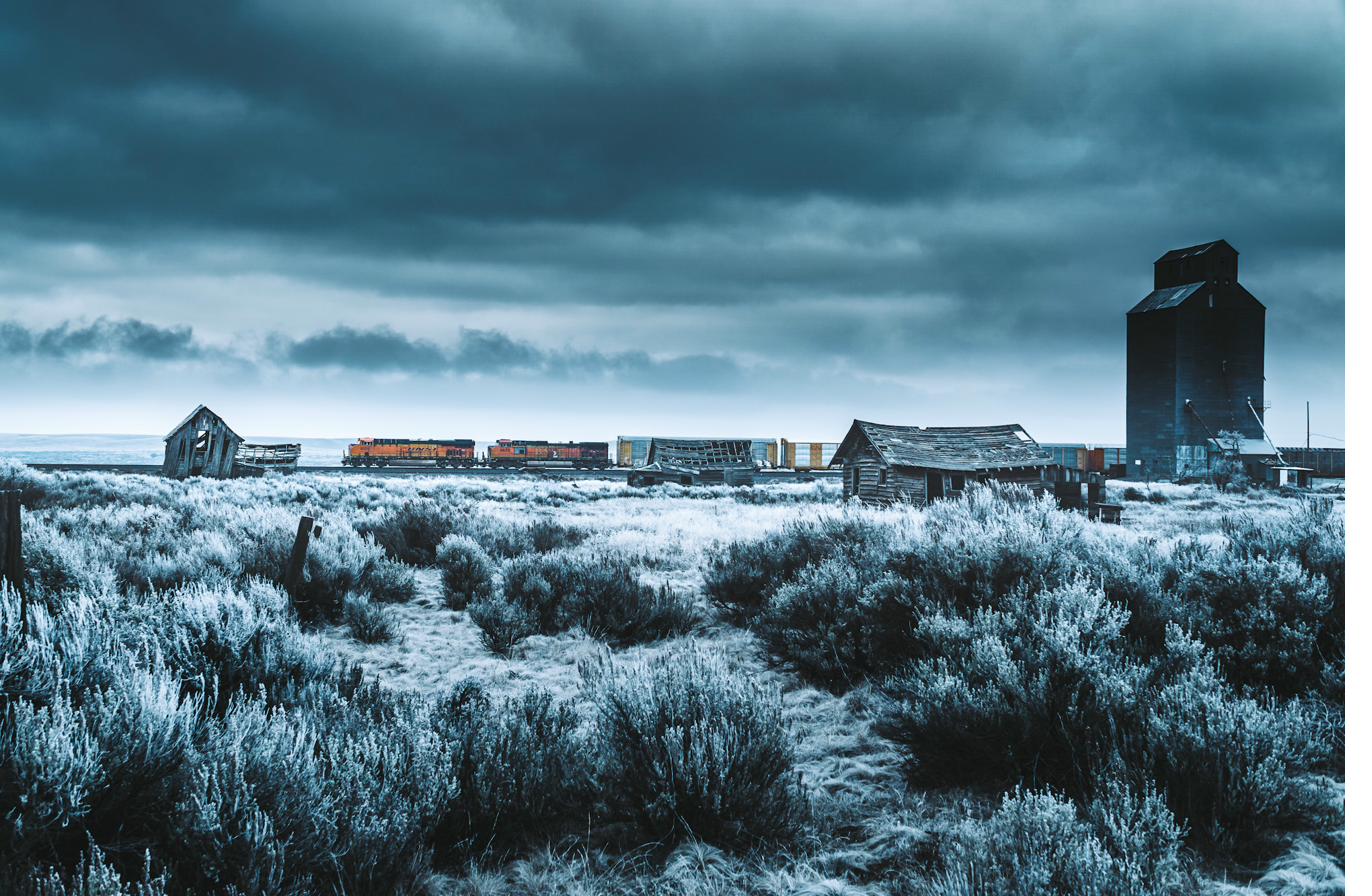BNSF freight threading abandoned homesteads through frost-white sagebrush under a bruised Eastern Washington sky